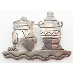 May include: Silver tone brooch depicting a person carrying a large, decorated pot. The person is wearing a hat and is standing on a wavy surface.