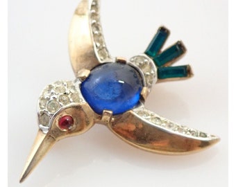 May include: A gold-tone hummingbird brooch with a blue cabochon stone, clear rhinestones, and green enamel accents.