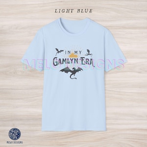 May include: Light blue t-shirt with a graphic design featuring a sunset, dragons, and the text "In My Gamlyn Era".