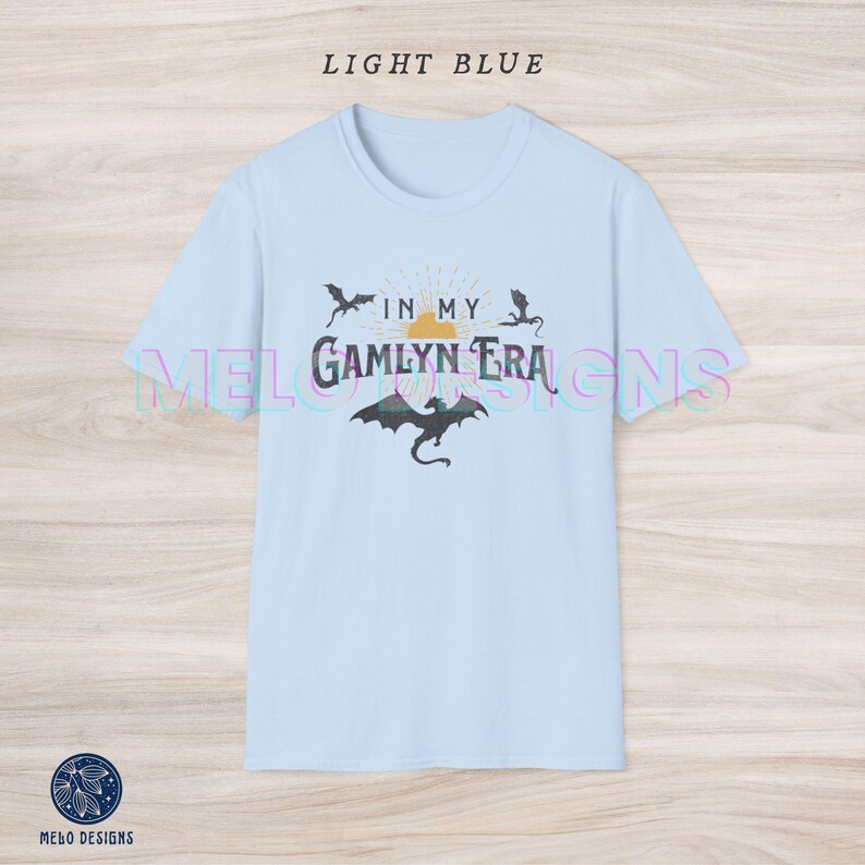 May include: Light blue t-shirt with a graphic design featuring a sunset, dragons, and the text "In My Gamlyn Era".