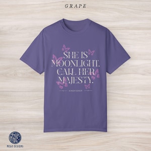May include: Purple t-shirt with a white graphic print that says "She is moonlight. Call her majesty." with a butterfly design and the word "Kingfisher" below the text.