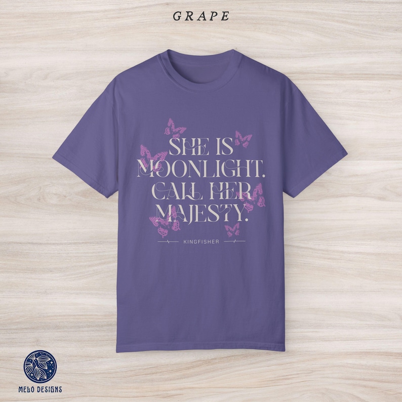 May include: Purple t-shirt with a white graphic print that says "She is moonlight. Call her majesty." with a butterfly design and the word "Kingfisher" below the text.