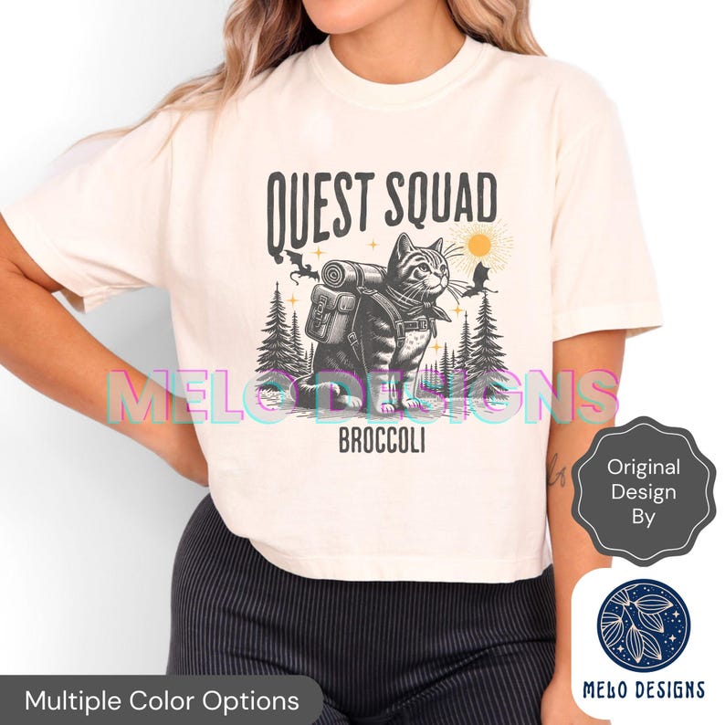 May include: A white t-shirt with a black and white illustration of a cat wearing a backpack and walking through a forest. The text "QUEST SQUAD" is above the cat and the text "BROCCOLI" is below the cat.