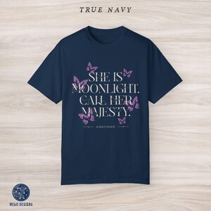 May include: A navy blue t-shirt with a white graphic print that says "She is moonlight. Call her majesty." with pink butterfly illustrations surrounding the text. The text "KINGFISHER" is printed below the graphic.