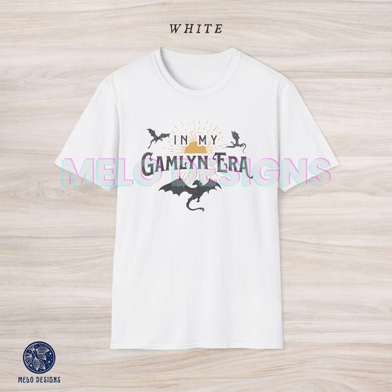 May include: White t-shirt with a graphic design featuring a sun with rays, three black dragons, and the text "In My Gamlyn Era".