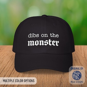 May include: Black baseball cap with the embroidered text "dibs on the monster" in white gothic lettering. The cap has a curved brim and a structured crown. The image also includes the text "Multiple Color Options" and a logo.