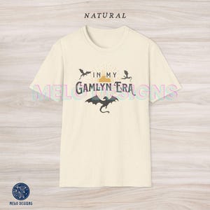 May include: A cream colored t-shirt with a graphic design featuring a sun with rays, three black dragons, and the text "In My Gamlyn Era".