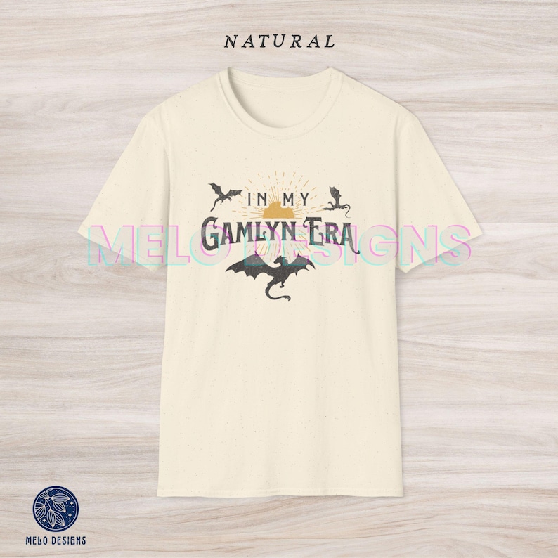 May include: A cream colored t-shirt with a graphic design featuring a sun with rays, three black dragons, and the text "In My Gamlyn Era".