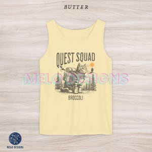 以下が含まれることがあります： バックパックを背負った猫のグラフィックと「QUEST SQUAD」と「BROCCOLI」の文字が印刷された黄色のタンクトップです。