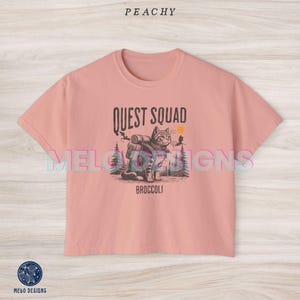 May include: Peachy pink cropped t-shirt with a graphic of a cat wearing a backpack and the text "Quest Squad Broccoli".