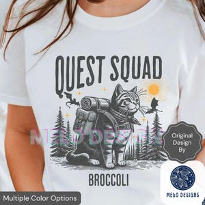 May include: White t-shirt featuring a graphic of a cat wearing a backpack, with the text "QUEST SQUAD" above and "BROCCOLI" below. The design includes trees, bats, and a sun.