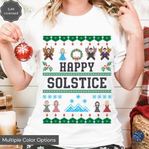 May include: White t-shirt with a green and red Christmas sweater design featuring the text "Happy Solstice". The design includes a wreath, snowflakes, mountains, and cartoon figures.