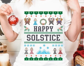 May include: White t-shirt with a green and red Christmas jumper design featuring the text "Happy Solstice". The design includes a wreath, snowflakes, mountains, and cartoon figures.