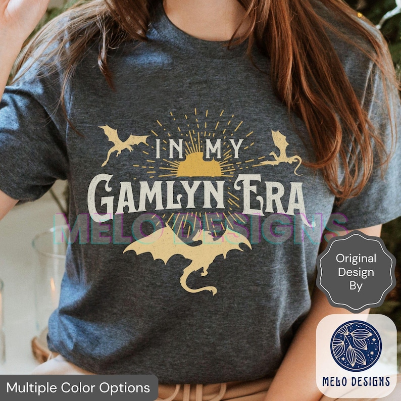 May include: A dark gray t-shirt with a graphic design featuring a sun, three dragons, and the text "In My Gamlyn Era".