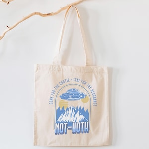 May include: Cream-colored tote bag with a blue and yellow graphic. The graphic features a UFO, mountains, and the text "COME FOR THE COOTIE - STAY FOR THE RESONANCE" and "NOT-HOTH". The bag has two handles and is hanging against a white wall.