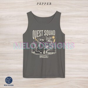 以下が含まれることがあります： バックパックを背負った猫のグラフィックと「QUEST SQUAD」と「BROCCOLI」のテキストがプリントされたグレーのタンクトップです。