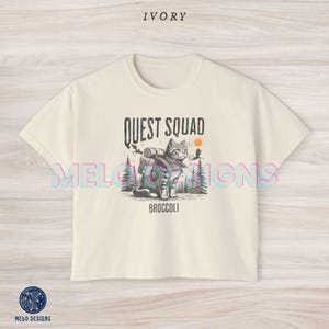 May include: A cream colored t-shirt with a graphic of a cat wearing a backpack and the text "Quest Squad Broccoli".