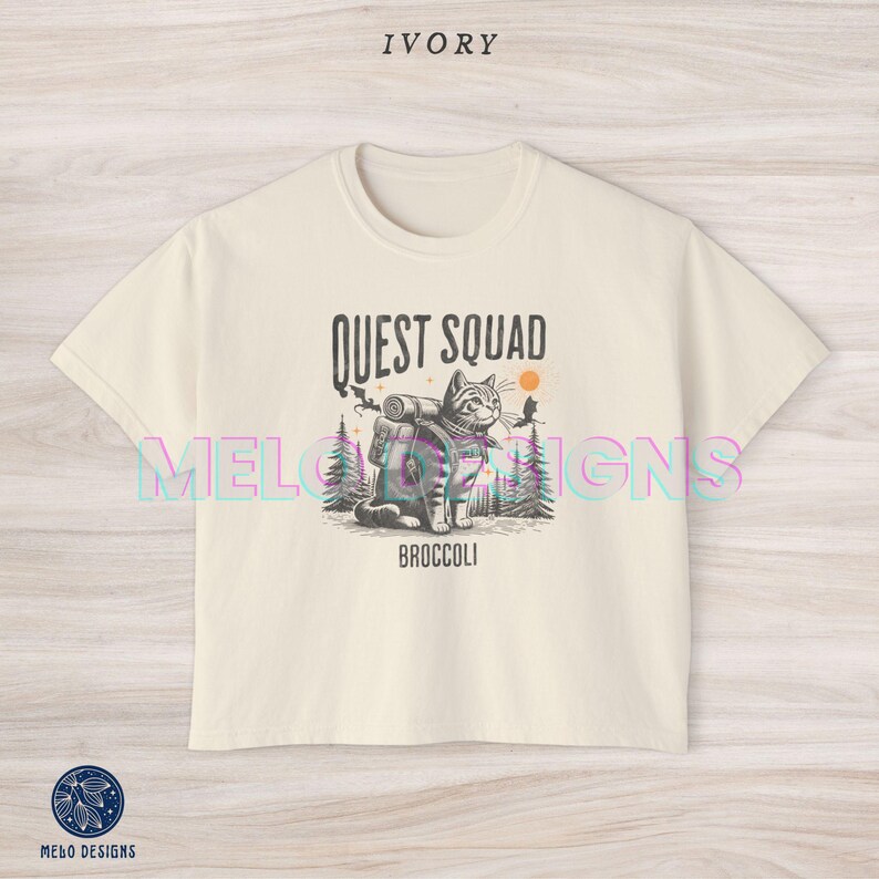 May include: A cream colored t-shirt with a graphic of a cat wearing a backpack and the text "Quest Squad Broccoli".