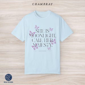 May include: Light blue t-shirt with a quote that reads "She is moonlight. Call her majesty." surrounded by butterfly graphics. The text "KINGFISHER" is printed below the quote.