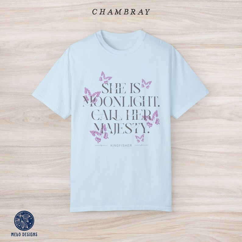 May include: Light blue t-shirt with a quote that reads "She is moonlight. Call her majesty." surrounded by butterfly graphics. The text "KINGFISHER" is printed below the quote.