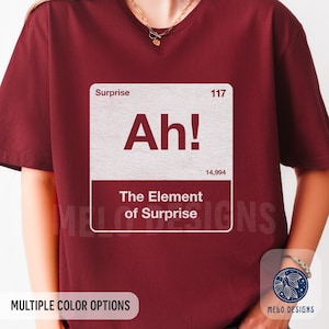 May include: Burgundy t-shirt with a white graphic that reads "Ah! The Element of Surprise." The design resembles a periodic table element box. The shirt also has the text "Multiple Color Options" and a logo.