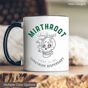 May include: White ceramic mug with a black handle. The mug has a green and black design with a skull smoking a cigarette and the text "Mirthroot Lunathion Light It Up Dispensary".