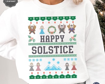 Puede incluir: Sudadera blanca con un diseño de solsticio de invierno verde, rojo y azul. El diseño presenta una corona, copos de nieve, montañas y personas. El texto "HAPPY SOLSTICE" está en el centro del diseño.