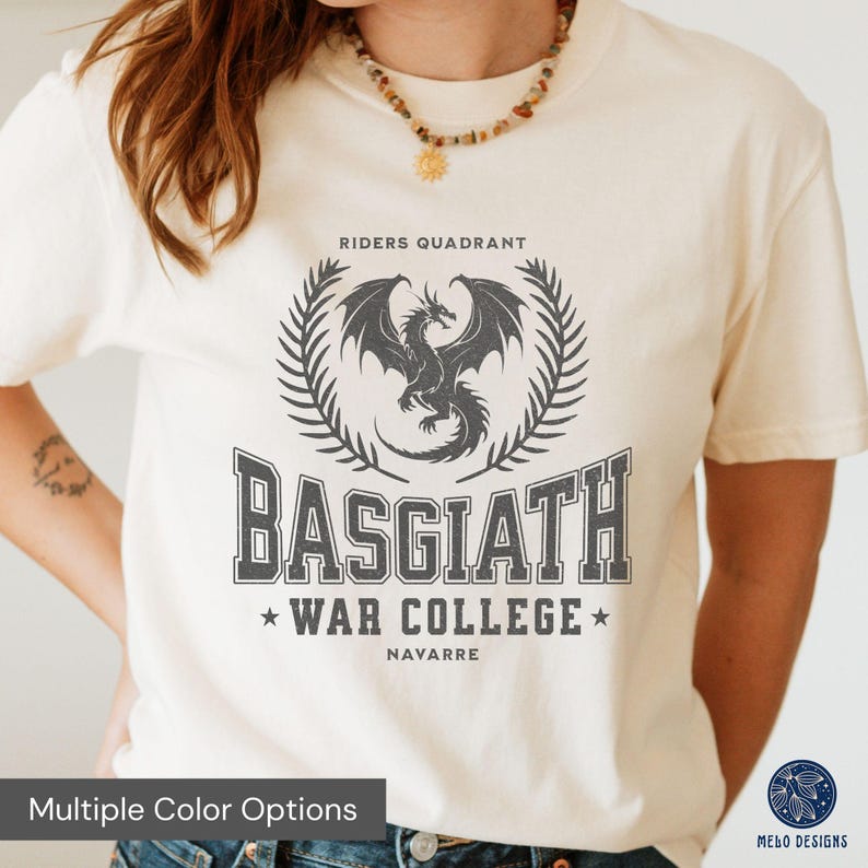 Puede incluir: Una camiseta blanca con un dise&ntilde;o gr&aacute;fico gris que presenta un drag&oacute;n, una corona de laurel y el texto "Riders Quadrant Basgiath War College Navarre".