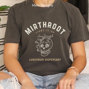 May include: A dark gray t-shirt with a white graphic of a skull smoking a cigarette. The text "Mirthroot Light It Up" is above the skull and "Lunathion Dispensary" is below the skull.