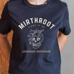 May include: A navy blue t-shirt with a white graphic of a skull smoking a cigarette. The text "Mirthroot Light It Up" is above the skull and "Lunathion Dispensary" is below the skull.