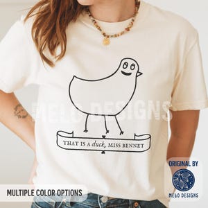 May include: A cream-colored short-sleeve t-shirt featuring a black line drawing of a duck with a smiling face. Below the duck is a banner graphic containing the text, THAT IS A DUCK, MISS BENNET, written in a classic serif font style.