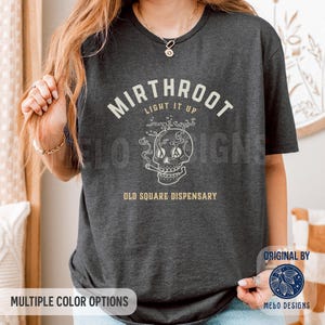 May include: A dark charcoal gray short-sleeve t-shirt featuring a white graphic of a skull with smoke, surrounded by the text MIRTHROOT, LIGHT IT UP, and OLD SQUARE DISPENSARY in a vintage-style font. The design is centered on the chest area.
