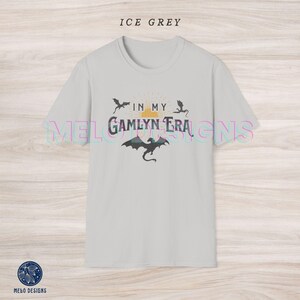 May include: A light grey t-shirt with a graphic design featuring a sunset, two dragons, and the text "In My Gamlyn Era".