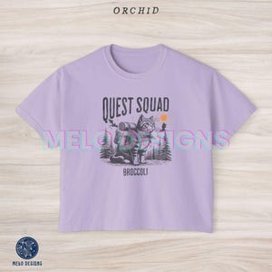 May include: A lavender colored cropped t-shirt with a graphic of a cat wearing a backpack and the text "Quest Squad" and "Broccoli".