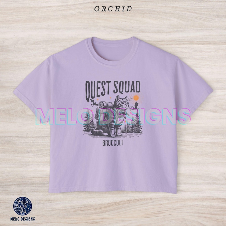 May include: A lavender colored cropped t-shirt with a graphic of a cat wearing a backpack and the text "Quest Squad" and "Broccoli".