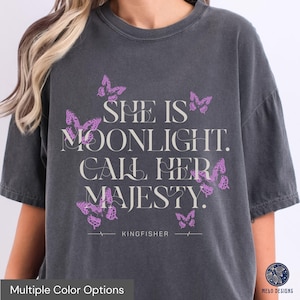 May include: A dark grey t-shirt with a graphic print of butterflies and the text "She is moonlight. Call her majesty." The text is in a light grey colour. The shirt also has the text "KINGFISHER" printed on the bottom.