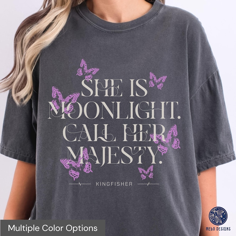May include: A dark grey t-shirt with a graphic print of butterflies and the text "She is moonlight. Call her majesty." The text is in a light grey colour. The shirt also has the text "KINGFISHER" printed on the bottom.