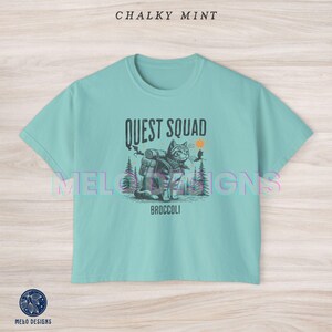 May include: A light blue t-shirt with a graphic of a cat wearing a backpack and the text "Quest Squad" and "Broccoli".