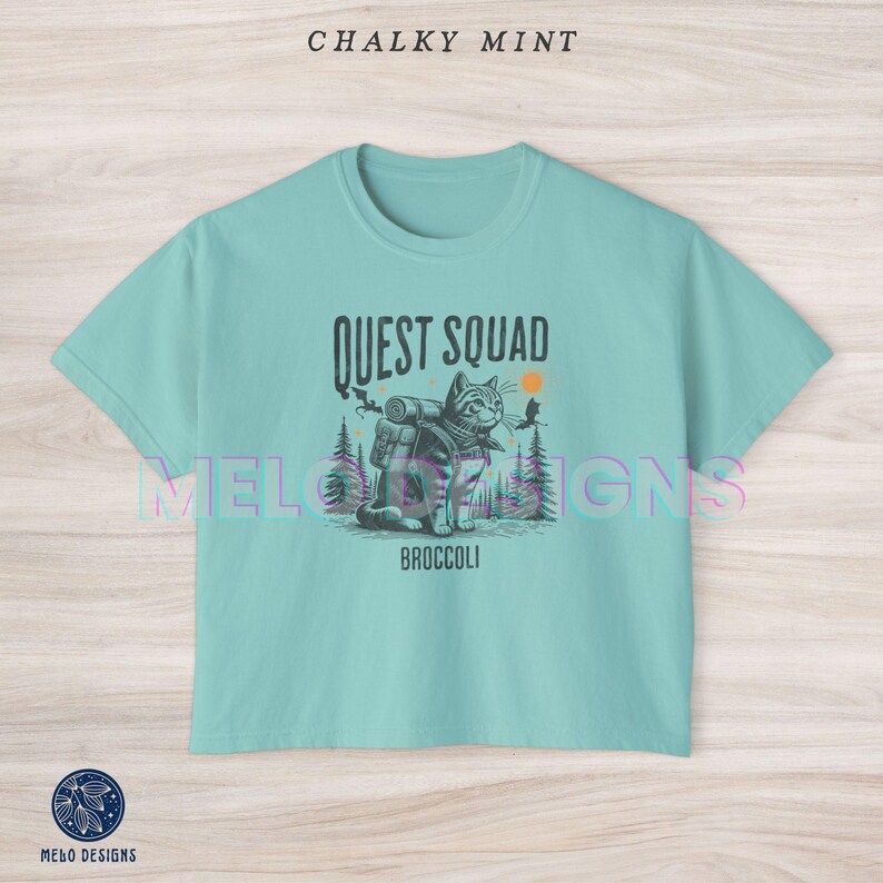 May include: A light blue t-shirt with a graphic of a cat wearing a backpack and the text "Quest Squad" and "Broccoli".