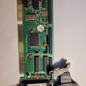 May include: A green printed circuit board featuring an LGS Prime 2C chip, a 24.000 MHz crystal oscillator, and various electronic components. The board includes a metal bracket with two ports and a sticker reading VOID IF REMOVED C/N 980326.