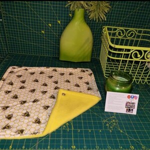 May include: A rectangular, patterned fabric with a bee and honeycomb design, paired with a yellow, triangular fabric. A green vase, a wire basket, and a candle sit nearby. A business card is also present.