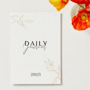 Op de afbeelding: Een wit dagboek met een gespikkelde textuur en gouden bladaccenten. De omslag heeft de tekst "DAILY Journal" en het jaar "2025".