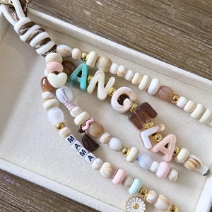 Peut inclure: Un porte-clés composé de trois brins de perles mélangées dans des tons crème, marron, rose et bleu. Les perles forment le nom ANGELA et le mot MAMA. Le design comprend une petite perle en forme de cœur, un pompon marron et une breloque fleur blanche.