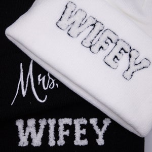May include: Two knitted beanies, one black and one white. The white beanie has the word "WIFEY" embroidered in a puffy, textured font. The black beanie has the word "WIFEY" and the word "Mrs." embroidered in a similar style.