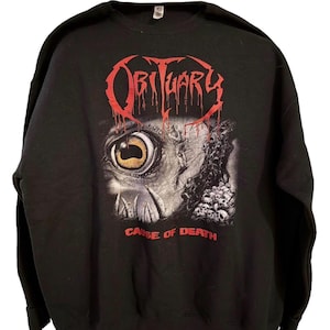 Puede incluir: Sudadera negra con el nombre de la banda "Obituary" en rojo goteante. El gráfico de abajo presenta un ojo de monstruo y calaveras. El texto "Cause of Death" está impreso en rojo debajo.