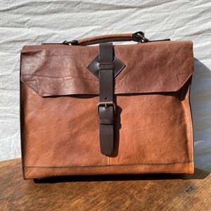 May include: A brown leather briefcase with a top handle and a front flap secured by a dark brown leather strap and buckle. The briefcase has a two-tone design, with a darker brown top section and a lighter brown body.