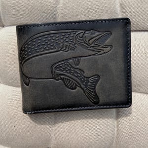 Può includere: Un portafoglio nero a due scomparti con un dettagliato design in rilievo di un pesce predatore con la bocca aperta e squame visibili. Il portafoglio presenta una finitura scura e liscia con cuciture visibili lungo i bordi del materiale.