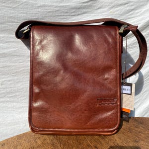 Op de afbeelding: Bruine leren messenger bag met een schouderriem. De tas is rechthoekig met een klepsluiting en een klein logo op de voorkant. De tas staat op een houten oppervlak.