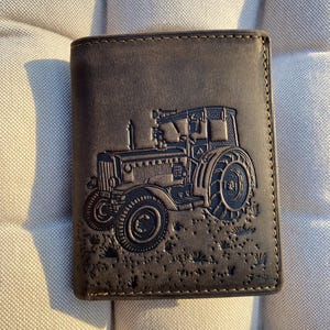 Op de afbeelding: Donkerbruine leren portemonnee met een gedetailleerde reliëf afbeelding van een vintage tractor. De portemonnee heeft een gestikte rand en een klassiek bi-fold ontwerp. De tractorafbeelding is in een donkerdere tint.