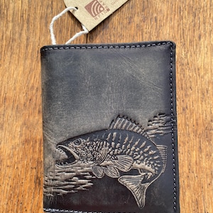 Puede incluir: Una cartera de cuero gris oscuro con un diseño de pez en relieve detallado. La cartera tiene un diseño de dos pliegues con costuras visibles y una etiqueta adjunta. La etiqueta tiene el texto "Jockey Club Germany".